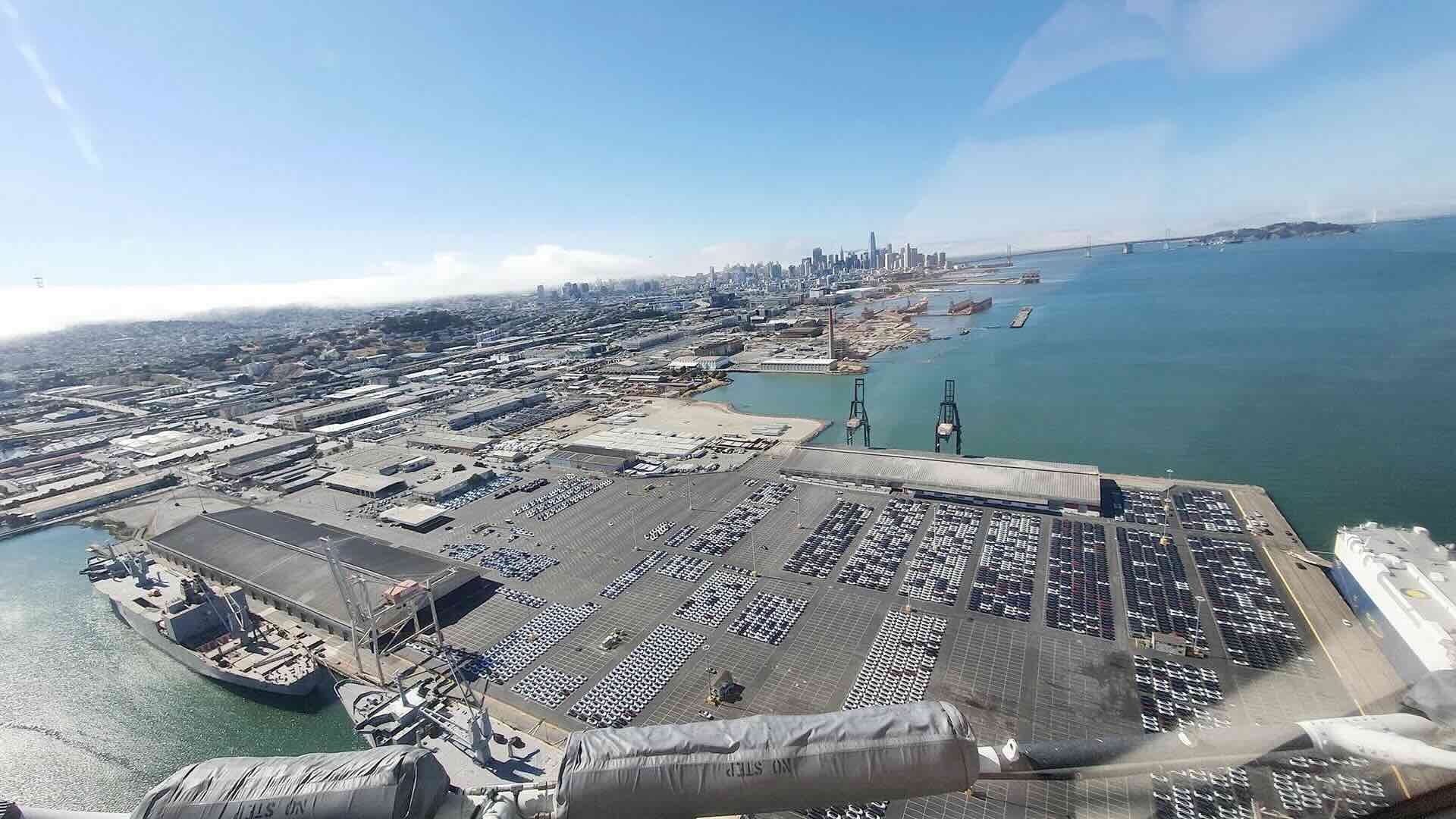 Aerial photograph of Pier 80, San Francisco, showing rows of imported automobiles staged on the apron, two gantry cranes, the warehouse transit shed, and downtown San Francisco in the background with fog rolling in from the west. A roll-on/roll-off vessel is moored at the right edge.